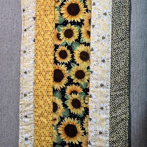 Puede incluir: Un edredón rectangular con un diseño de patchwork. El edredón presenta paneles de girasoles sobre negro, un patrón de panal, flores blancas con abejas y un patrón de lunares negros y amarillos. El edredón es principalmente amarillo, negro y blanco.