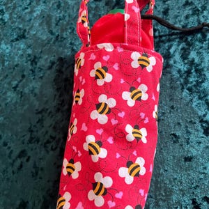 Può includere: Una borsa in tessuto rosso con un motivo ripetuto di fiori bianchi e api gialle e nere. La borsa ha un manico abbinato e una chiusura con coulisse. L'interno è rosso. La borsa è progettata per contenere una bottiglia.