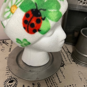 Puede incluir: Gorro de forro polar blanco con estampado de mariquita, tréboles verdes y margaritas. El gorro está adornado con una mariquita roja con lunares negros. Los tréboles y las margaritas son verdes y amarillas.