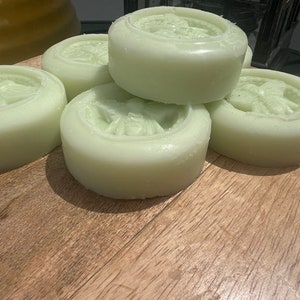 May include: Five round, pale green soap bars with a raised design of a bee on the top of each bar. The bars are stacked on top of each other on a wooden surface.