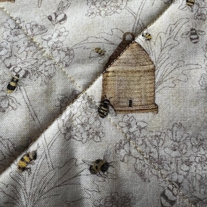May include: Close-up of a quilted fabric with a bee and beehive pattern. The fabric is beige with a floral design in gray and brown. Bees are scattered throughout the design, with a beehive in the center.