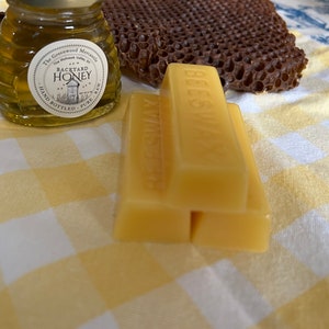 May include: Three yellow beeswax blocks stacked on top of each other on a yellow and white checked tablecloth. The top block has the word "Beeswax" printed on it. A small glass jar of honey with a label that reads "Backyard Honey" is in the background.