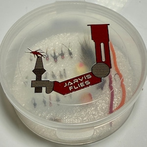 May include: A clear plastic container filled with various fishing flies. The container has a red and brown graphic that reads "JARVIS FLIES". The flies are in different colors and sizes, with some having feathers and other materials.