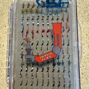 May include: A clear plastic fly fishing tackle box containing an assortment of small artificial flies arranged in rows on a grey foam insert. The lid features a blue SF logo and a large orange graphic of a fly tying vise with the text Jarvis Flies.