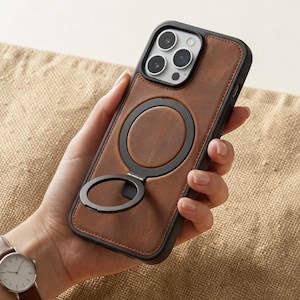 May include: Brown leather phone case with a black frame and a circular ring holder. The case has a stitched border and a black ring that swivels out for use as a stand. The phone case is designed to protect the phone.