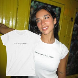 以下が含まれることがあります： 「Show me your books」のテキストが入った白いTシャツ。Tシャツは半袖で、クルーネックです。テキストはシンプルなサンセリフ体です。Tシャツは人の前に掲げられています。
