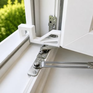 Può includere: Primo piano di un telaio di finestra bianco con una cerniera metallica e un fermo di plastica bianco. La cerniera è argentata e fissata con viti. Lo sfondo è una vista sfocata di fogliame verde.