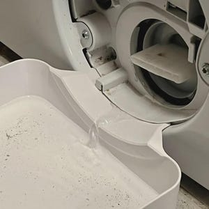 Pode incluir: Close-up do compartimento do filtro de drenagem de uma máquina de lavar roupa branca. Uma bandeja branca está posicionada abaixo da abertura, recolhendo água. O compartimento do filtro está aberto, revelando o filtro e os componentes internos.