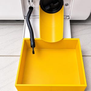 Op de afbeelding: Een wit onderhoudspaneel van een apparaat staat open en toont een zwart rond afvoerfilter, een zwarte flexibele afvoerslang en een felgele kunststof afvoergoot. Een vierkante gele opvangbak is onder de goot geplaatst.
