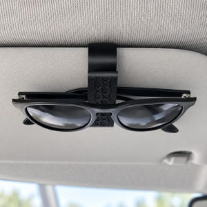 May include: A black plastic car visor clip featuring a textured circular pattern on the front surface. The clip is securely attached to a light grey vehicle sun visor and holds a pair of black-framed sunglasses in a horizontal position.