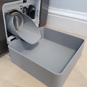 May include: A grey plastic washing machine drain filter assembly, with a matching grey rectangular tray beneath. The filter is open, showing internal parts. The setup is on a tiled floor.