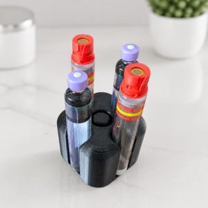 Może przedstawiać: Czarny, wydrukowany w technologii 3D plastikowy stojak z czterema cylindrycznymi otworami, w których umieszczono cztery peny insulinowe. Dwa peny mają jaskrawoczerwone zatyczki z żółtymi paskami, a pozostałe dwa mają jasnofioletowe zatyczki.