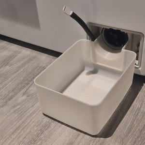 Può includere: Un contenitore rettangolare di plastica bianca posizionato sotto l'uscita di scarico di una lavatrice. Un tubo nero si estende dalla macchina al contenitore. Il contenitore è su un pavimento di legno.