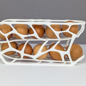 Może przedstawiać: Biały, dwupoziomowy pojemnik na jajka wypełniony brązowymi jajkami. Pojemnik ma otwartą, organiczną konstrukcję, umożliwiającą widoczność jajek. Jajka są mieszanką jasnych i ciemnobrązowych. Pojemnik stoi na białej powierzchni.