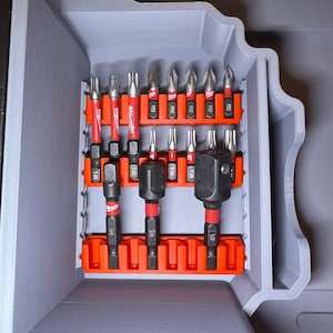 Puede incluir: Una caja de herramientas gris con un inserto naranja que contiene varias puntas de destornillador negras y rojas. Las puntas están etiquetadas con tamaños y tipos, incluyendo T-25, 1/4, 3/8 y 1/2 pulgadas, y tipos PH2, PZ3 y T-20.