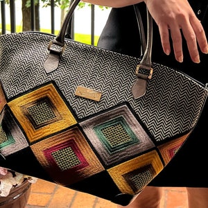 May include: A black and white chevron patterned tote bag with a geometric design featuring squares in shades of yellow, red, green, and brown. The bag has a gold metal logo that says "Handmade by"