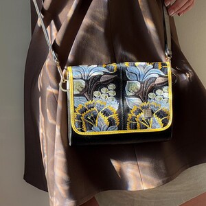 May include: A black crossbody bag with a floral pattern in blue, yellow, and white. The bag has a gold trim and a silver strap. The bag is rectangular and appears to be made of a textured material.