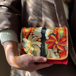 May include: A vibrant, embroidered clutch bag with a red base and a black central panel. It features colourful floral designs in orange, yellow, and green. The bag has a gold clasp and a tan trim.