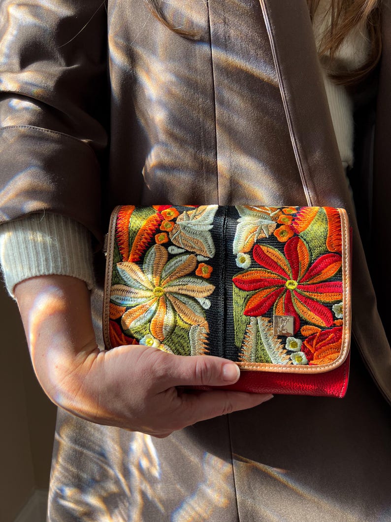 May include: A vibrant, embroidered clutch bag with a red base and a black central panel. It features colourful floral designs in orange, yellow, and green. The bag has a gold clasp and a tan trim.