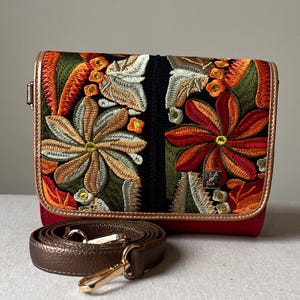 May include: A red crossbody bag with an embroidered floral design in orange, green, and white. The bag has a brown strap and gold-coloured hardware. The bag's edges are trimmed with a metallic gold material.