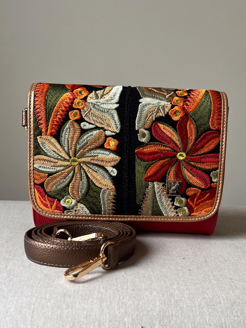 May include: A red crossbody bag with an embroidered floral design in orange, green, and white. The bag has a brown strap and gold-coloured hardware. The bag's edges are trimmed with a metallic gold material.
