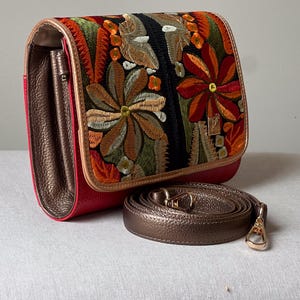 May include: A red and bronze crossbody bag with an embroidered floral design. The bag features a bronze strap and a gold-coloured clasp. The flap of the bag is decorated with colourful embroidered flowers.