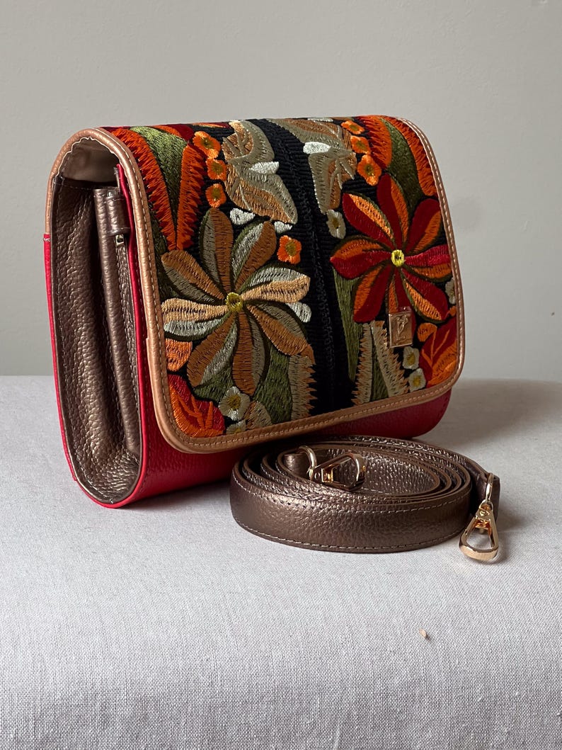 May include: A red and bronze crossbody bag with an embroidered floral design. The bag features a bronze strap and a gold-coloured clasp. The flap of the bag is decorated with colourful embroidered flowers.