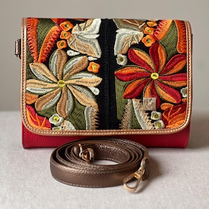 May include: A red leather crossbody bag with a flap closure. The flap features vibrant floral embroidery in orange, green, and cream. A detachable bronze strap and gold-tone hardware are also visible.