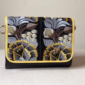 May include: Black clutch with embroidered floral design in gray, gold, and brown. The clutch has a yellow trim and a metal ring on the side. The floral pattern is on the front flap.