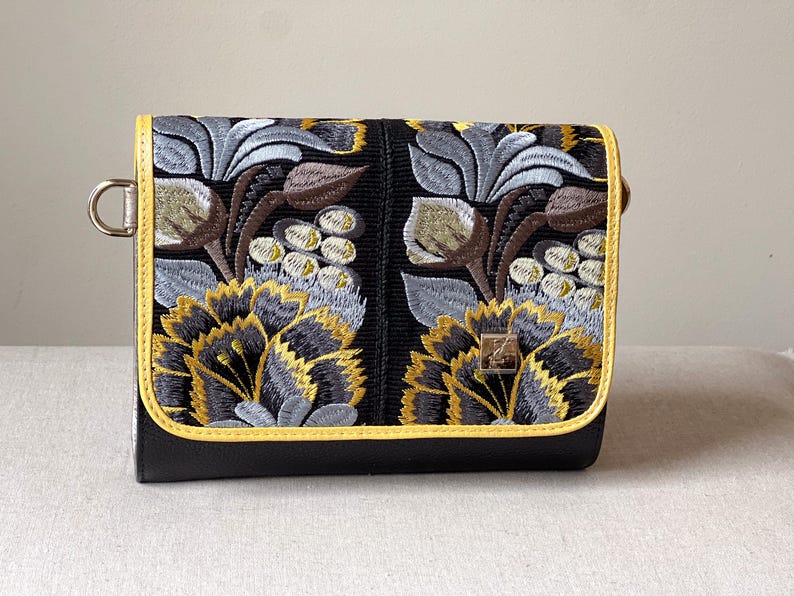 May include: Black clutch with embroidered floral design in gray, gold, and brown. The clutch has a yellow trim and a metal ring on the side. The floral pattern is on the front flap.