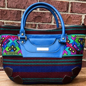 May include: A blue handbag with a colorful woven design. The bag features a blue leather handle and a front pocket. The woven pattern includes geometric shapes in pink, green, and purple. The bottom corners are brown.