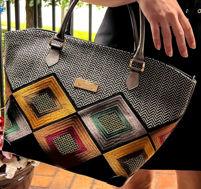 May include: A black and white chevron patterned tote bag with a geometric design featuring squares in shades of yellow, red, green, and brown. The bag has a gold metal logo that says "Handmade by"