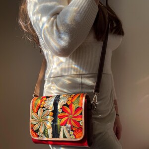 May include: A crossbody bag with a colourful floral design. The bag has a red base, a flap with embroidered flowers in orange, yellow, and green, and a brown leather strap. The bag has a silver clasp and a brown leather trim.