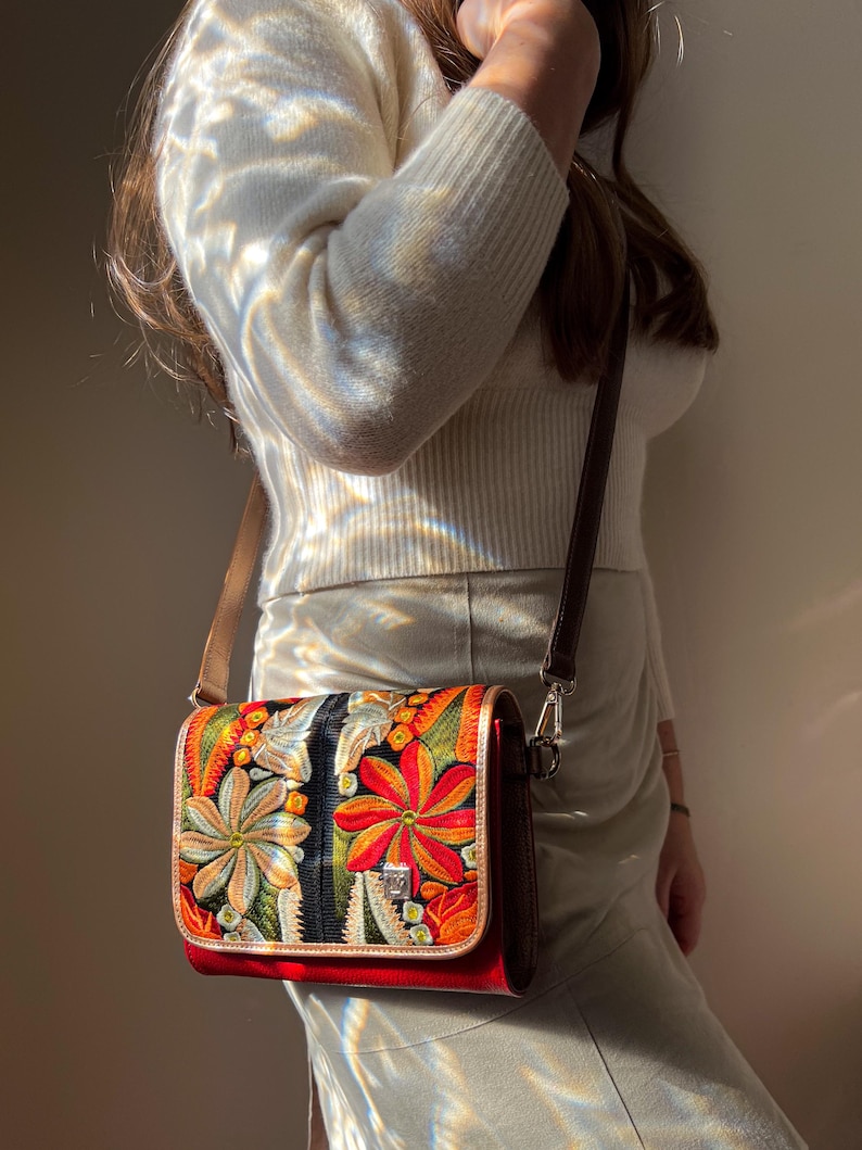 May include: A crossbody bag with a colourful floral design. The bag has a red base, a flap with embroidered flowers in orange, yellow, and green, and a brown leather strap. The bag has a silver clasp and a brown leather trim.