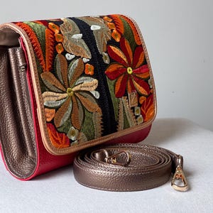 May include: A red handbag with an embroidered floral design in orange, green, and brown. The bag has a bronze-coloured strap and interior. The clasp is gold-coloured. The bag is on a white surface.
