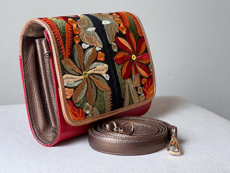 May include: A red handbag with an embroidered floral design in orange, green, and brown. The bag has a bronze-coloured strap and interior. The clasp is gold-coloured. The bag is on a white surface.