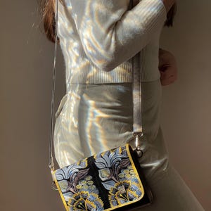 May include: A black crossbody bag with a floral design in blue, yellow, and silver. The bag has a gold trim and a silver strap. The bag is rectangular and appears to be made of a textured fabric.