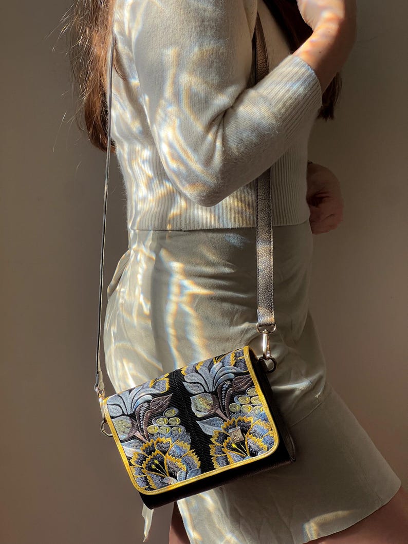 May include: A black crossbody bag with a floral design in blue, yellow, and silver. The bag has a gold trim and a silver strap. The bag is rectangular and appears to be made of a textured fabric.