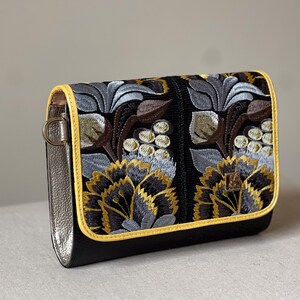 May include: Black clutch with embroidered floral design in gold, gray, and brown. The bag has a yellow trim and silver-colored sides. The floral pattern is detailed and covers the front of the bag.