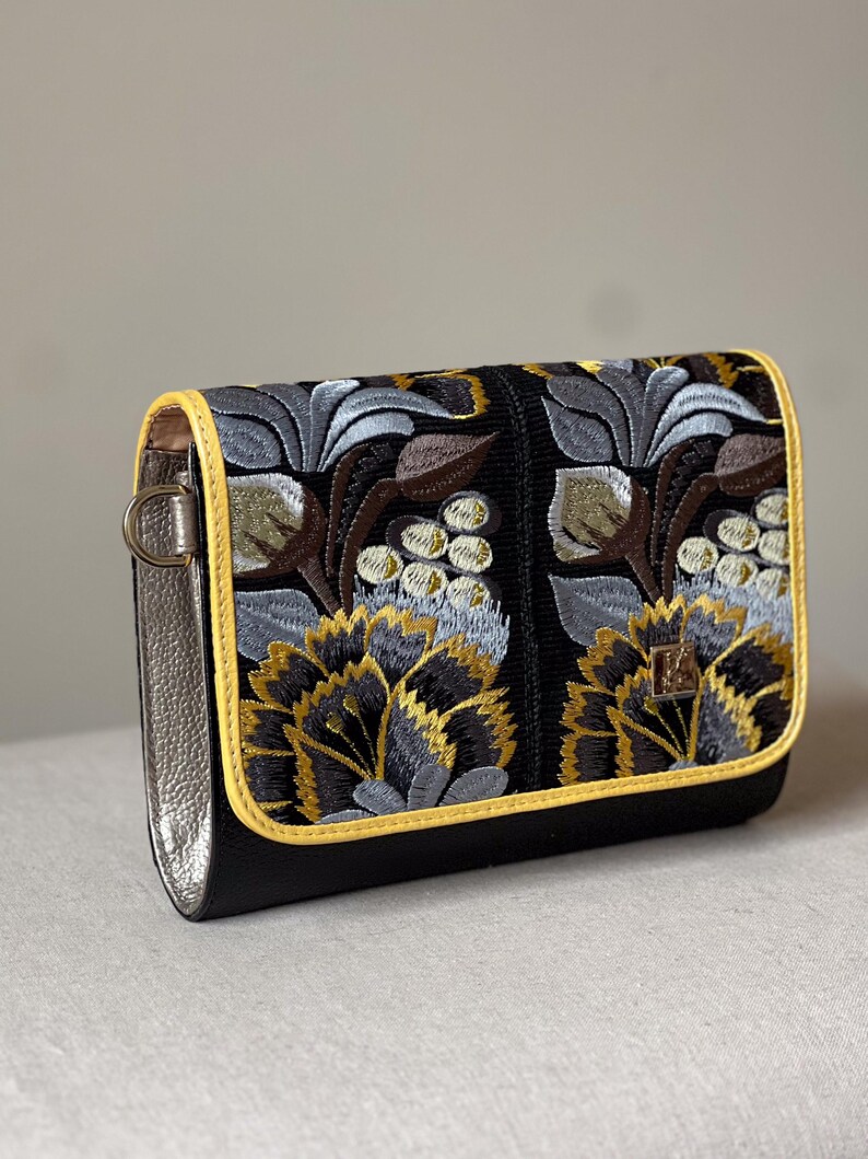 May include: Black clutch with embroidered floral design in gold, gray, and brown. The bag has a yellow trim and silver-colored sides. The floral pattern is detailed and covers the front of the bag.