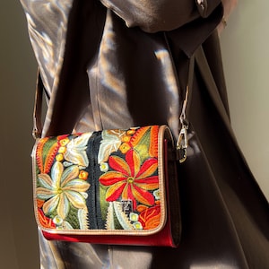 May include: A rectangular crossbody bag with a vibrant floral embroidery design. The bag features a red base, a black central panel, and a tan trim. The strap is a light brown colour, and the bag has a silver clasp.