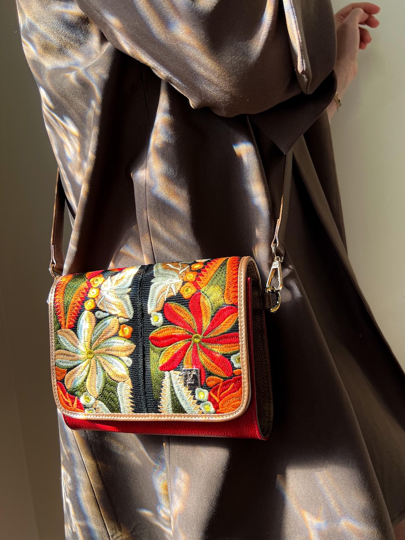 May include: A rectangular crossbody bag with a vibrant floral embroidery design. The bag features a red base, a black central panel, and a tan trim. The strap is a light brown colour, and the bag has a silver clasp.
