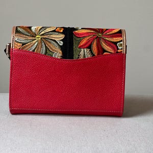 May include: A red handbag with a flap featuring embroidered floral designs in shades of orange, yellow, and green on a black background. The bag has a textured red front and a gold-coloured trim.