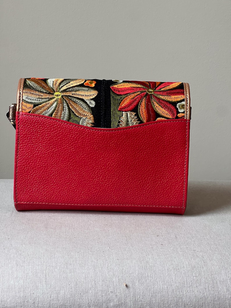 May include: A red handbag with a flap featuring embroidered floral designs in shades of orange, yellow, and green on a black background. The bag has a textured red front and a gold-coloured trim.