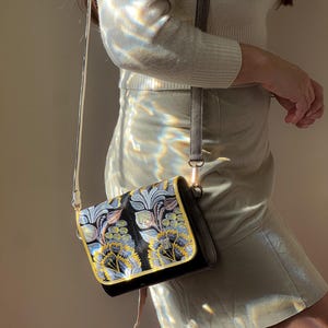 May include: A black crossbody bag with a floral design in blue, yellow, and gray. The bag has a gold trim and a gray strap. The bag is made of a textured material and has a rectangular shape.