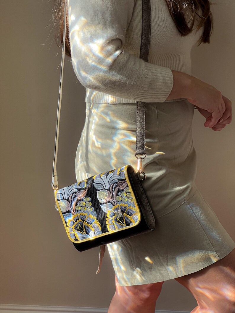 May include: A black crossbody bag with a floral design in blue, yellow, and gray. The bag has a gold trim and a gray strap. The bag is made of a textured material and has a rectangular shape.