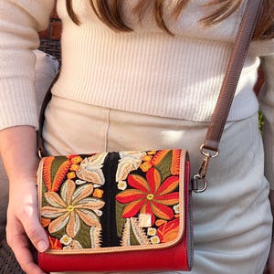 May include: A red crossbody bag with a colourful floral embroidered flap. The bag has a brown leather strap and a gold-coloured clasp. The embroidered design includes orange, yellow, and green flowers.