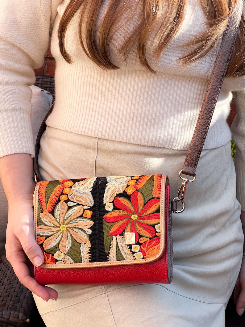 May include: A red crossbody bag with a colourful floral embroidered flap. The bag has a brown leather strap and a gold-coloured clasp. The embroidered design includes orange, yellow, and green flowers.