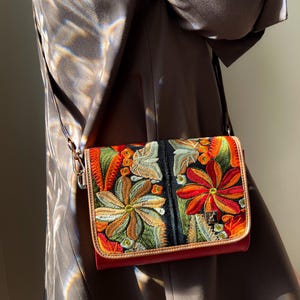 May include: A crossbody bag with colourful floral embroidery. The bag has a red base, a black central panel, and a tan border. The embroidery features orange, green, and yellow flowers. It has a long, thin strap.