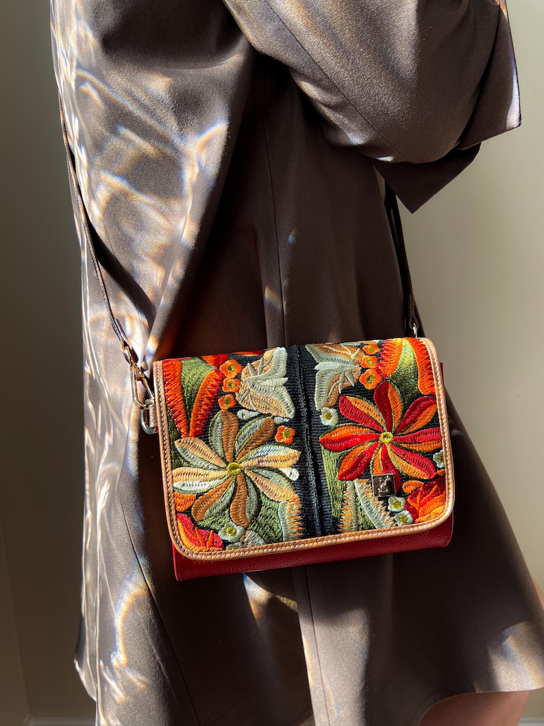May include: A crossbody bag with colourful floral embroidery. The bag has a red base, a black central panel, and a tan border. The embroidery features orange, green, and yellow flowers. It has a long, thin strap.